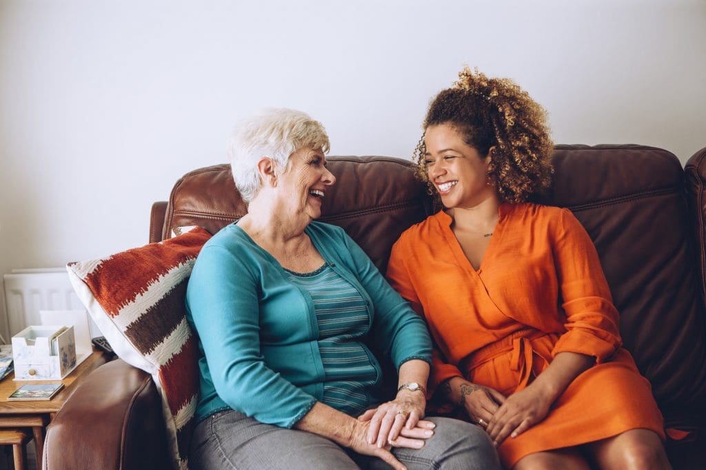 A home carer visiting a lady in her home, sat together on the sofa, smiling at one another. Aster Care are proud of the care they provide.